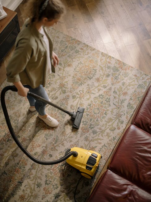 A woman using a yellow vacuum cleaner to deep clean a patterned area rug with floral motifs in a living room setting, which features wooden flooring and a leather sofa. The room is well-lit, and the r