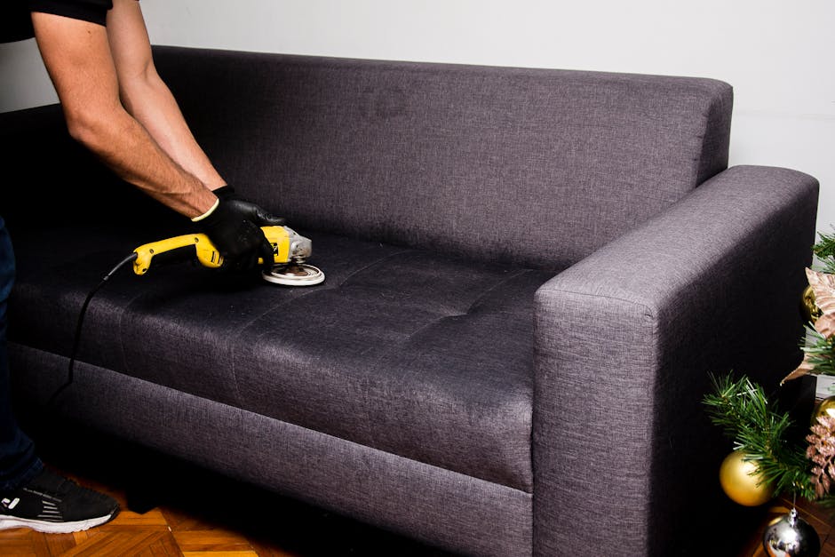 A person wearing black gloves is using a handheld electric cleaning device to deep clean a dark gray fabric sofa in a residential living room. The sofa has a modern design with wide armrests and a plu