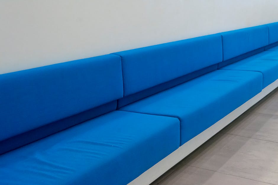 A person wearing black gloves is using a handheld electric cleaning device to deep clean a dark gray fabric sofa in a residential living room. The sofa has a modern design with wide armrests and a plu