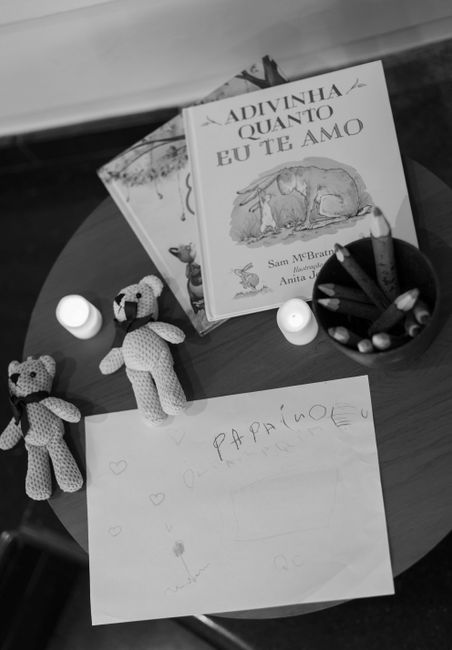Black and white image showing a wooden table with children's drawings and toys. On the table, there are two small knitted teddy bears, a sheet of paper with hearts and the handwritten word 'papá' in S